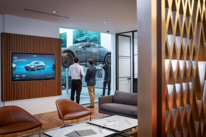 Car dealership showroom featuring two men looking at a display car and a TV screen showing an Aston Martin DBX model.