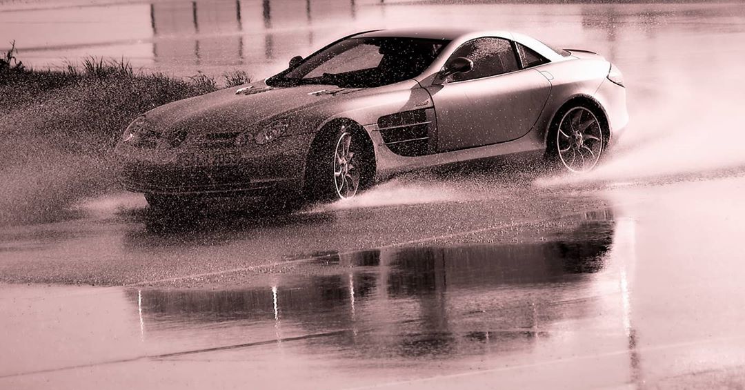 Silver Mercedes-Benz SLR McLaren driving on wet asphalt, reflections visible, near grassy area, showcasing automotive performance and design.