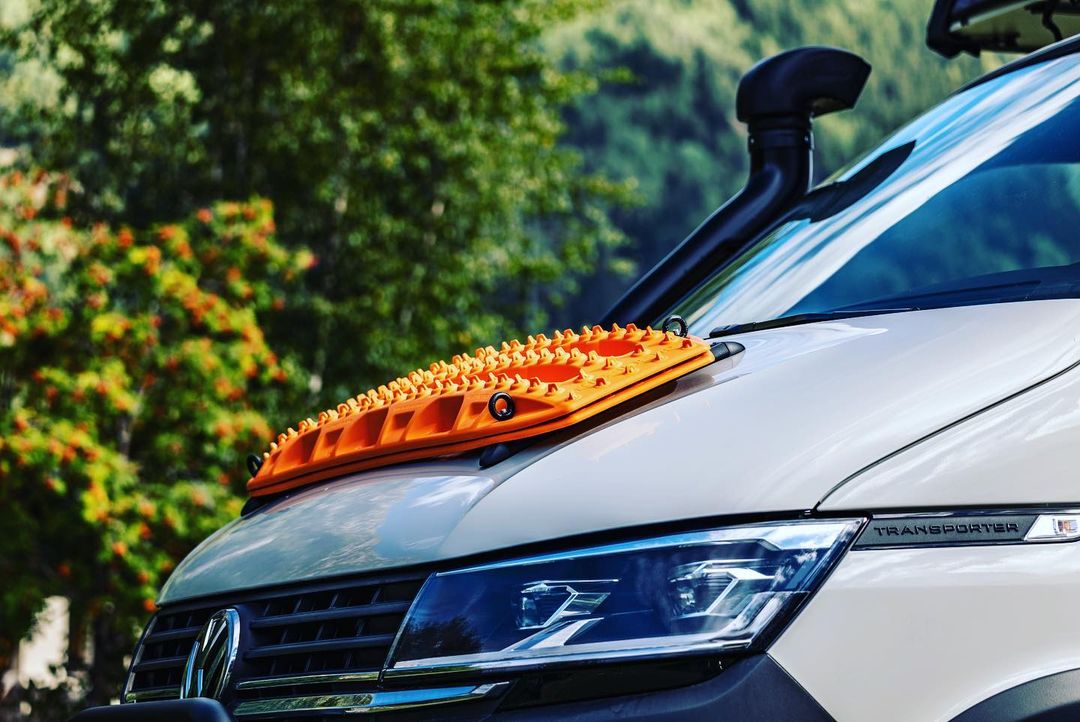 Volkswagen Transporter van with an orange traction board mounted on the hood, parked outdoors near leafy green trees and grass.