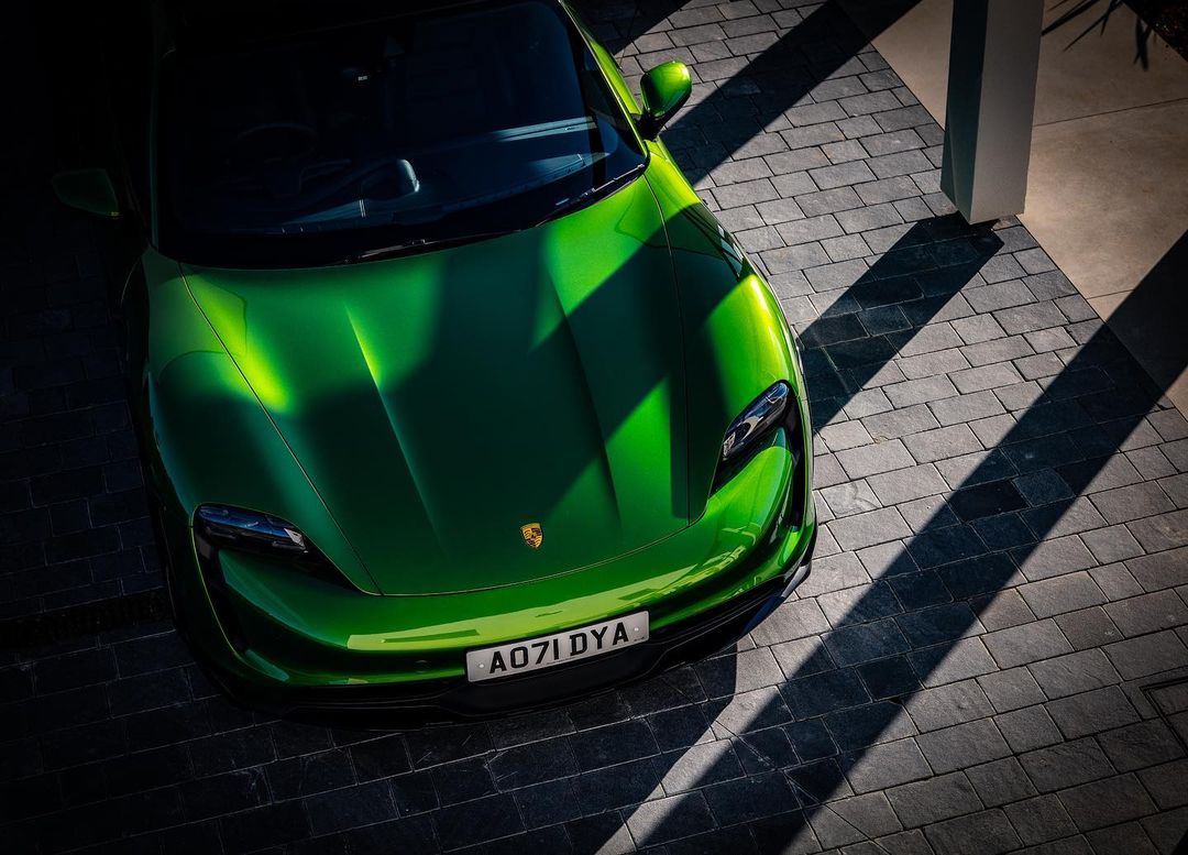 Overhead shot of a bright green Porsche Taycan, parked on a patterned stone driveway, with strong shadows and dappled sunlight.