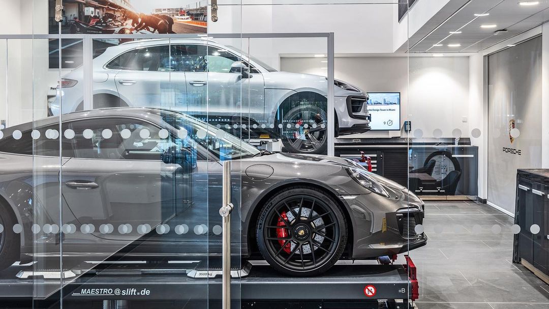 Two gray Porsche cars, one on a lift in the foreground, and one in the background, inside of a dealership.