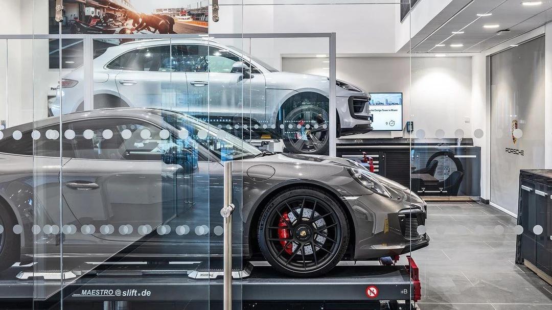 Two gray Porsche cars, one on a lift in the foreground, and one in the background, inside of a dealership.