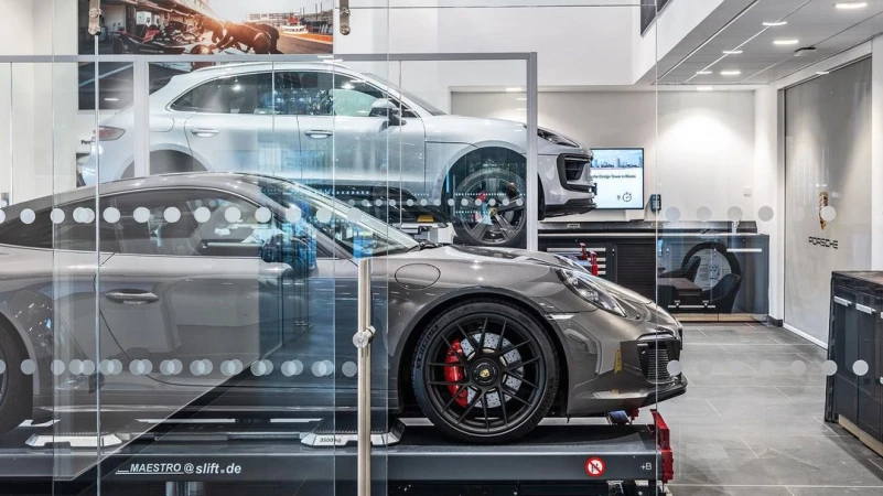 Two gray Porsche cars, one on a lift in the foreground, and one in the background, inside of a dealership.