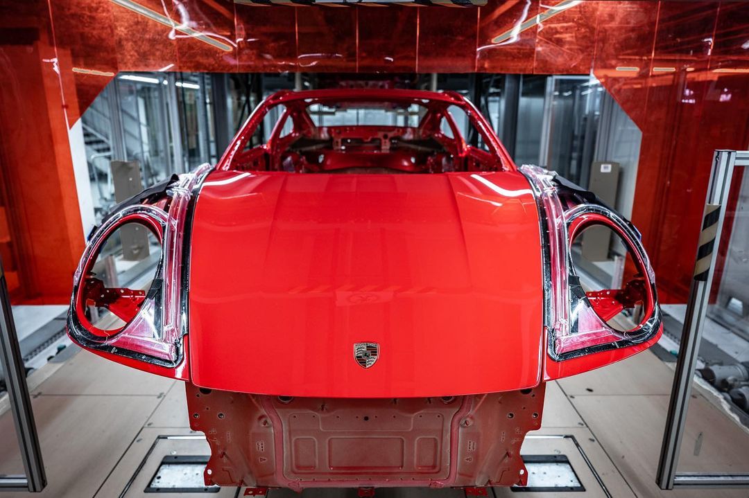 Close-up of a bright red Porsche chassis with no wheels or windows in a factory setting; the company logo is centered on the hood.