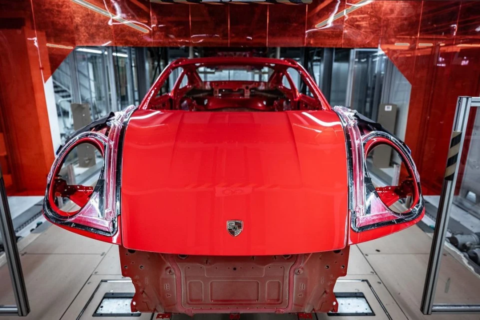 Close-up of a bright red Porsche chassis with no wheels or windows in a factory setting; the company logo is centered on the hood.