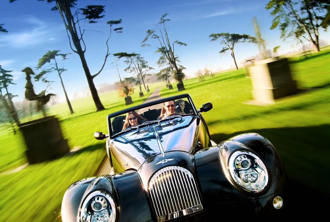 Two adults in a vintage black convertible car driving on a green grass-lined road with trees under a blue sky on a sunny day.