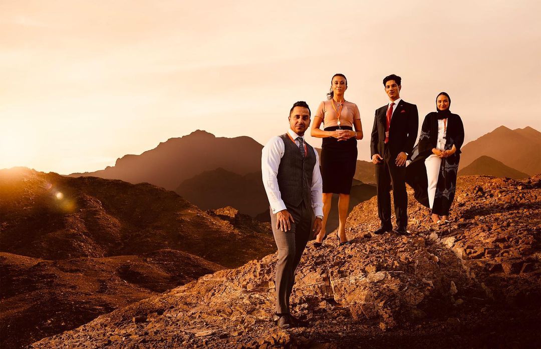 Four businesspeople stand on a rocky hilltop with mountains in the background at sunset, dressed in formal attire.