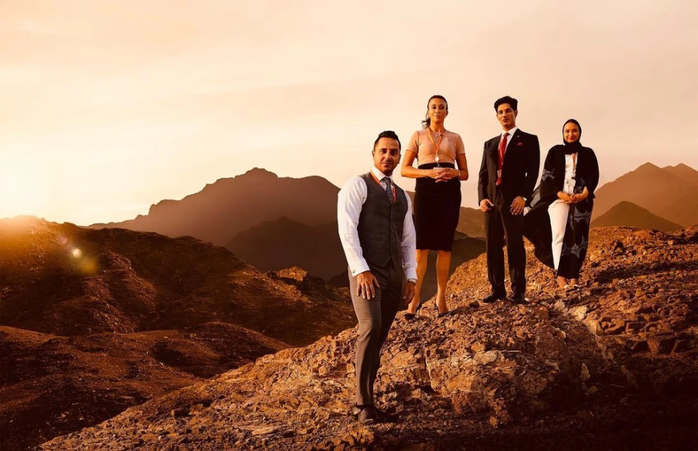 Four businesspeople stand on a rocky hilltop with mountains in the background at sunset, dressed in formal attire.