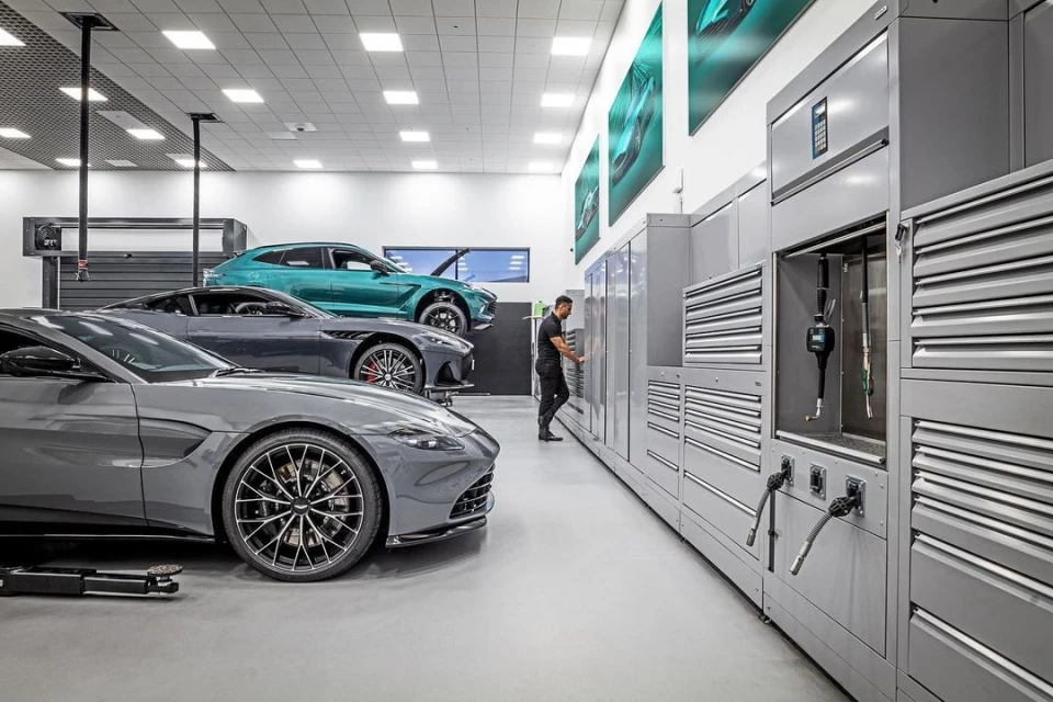 Three gray and green Aston Martin vehicles are displayed in a garage with an auto mechanic working near gray metal storage units.