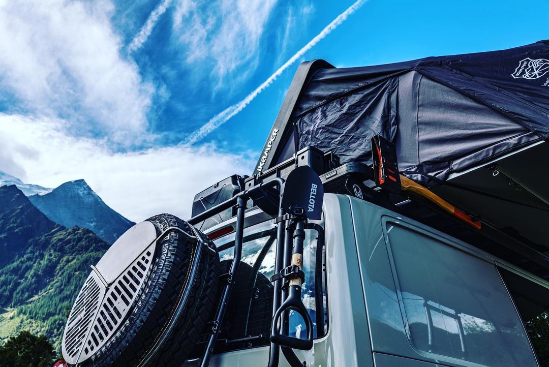Silver campervan with a rooftop tent, ladder, and spare tire against a backdrop of a blue sky, clouds, and distant mountains.