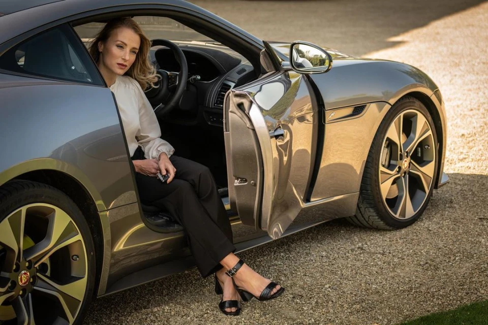 A woman sits in a gray Jaguar F-Type on a gravel drive. She wears black pants, heeled sandals, and a cream blouse.