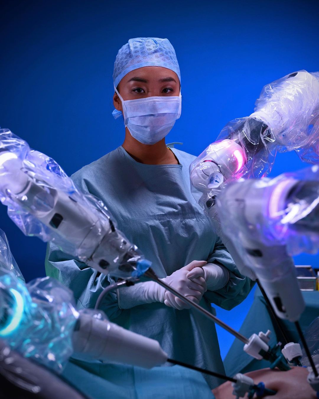 Medical professional in surgical scrubs and face mask stands next to robotic surgery equipment in a blue operating room environment.
