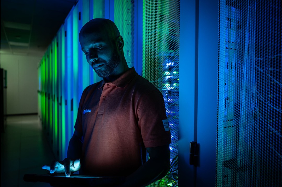 Man works in a data center, interacting with server racks illuminated by blue and green lights. He is wearing a polo shirt.