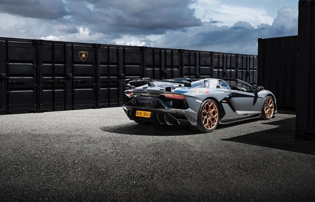 Gray Lamborghini Aventador SVJ Roadster parked near black shipping containers under a cloudy sky, showcasing automotive design and luxury.