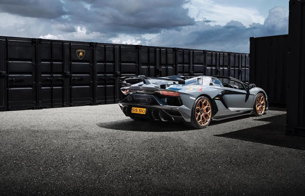 Gray Lamborghini Aventador SVJ Roadster parked near black shipping containers under a cloudy sky, showcasing automotive design and luxury.