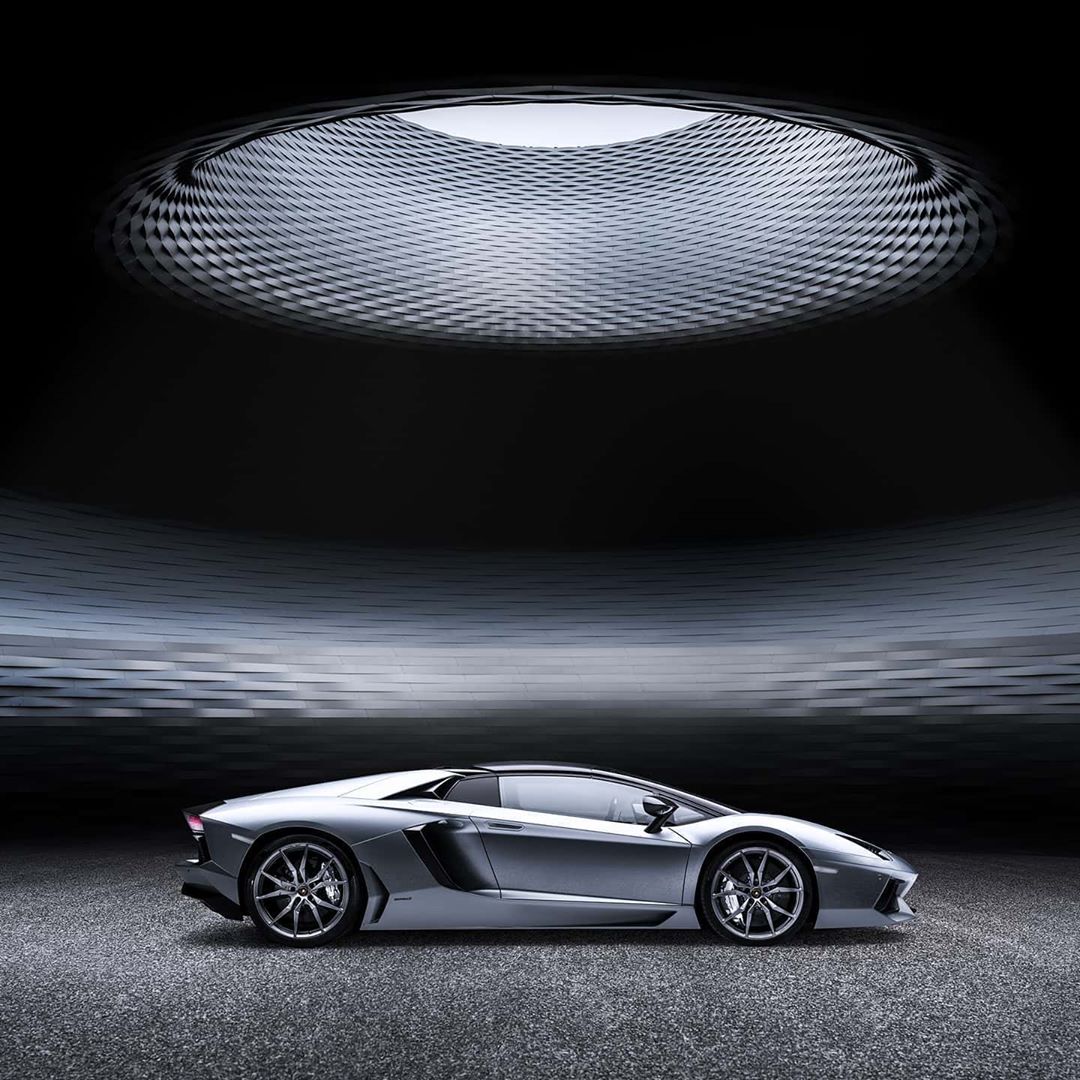Silver Lamborghini Aventador in a modern showroom with textured silver walls and ceiling under a large skylight, capturing sleek automotive design.