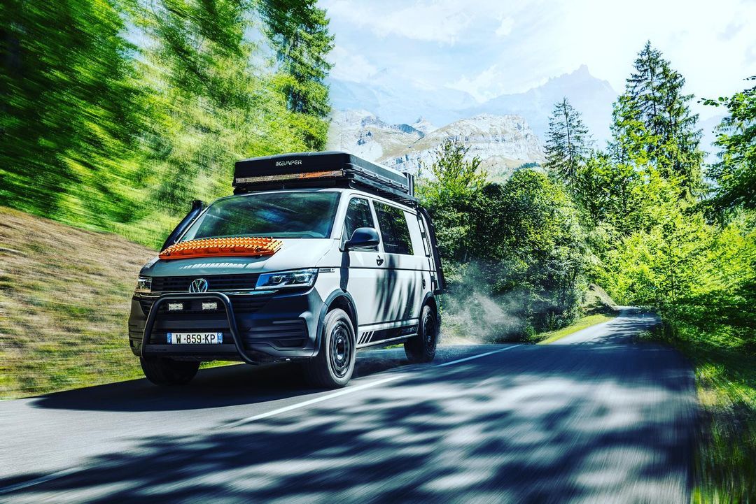 White Volkswagen van with a roof rack and off-road tires driving on a paved road through a bright green forest with mountains in the distance.