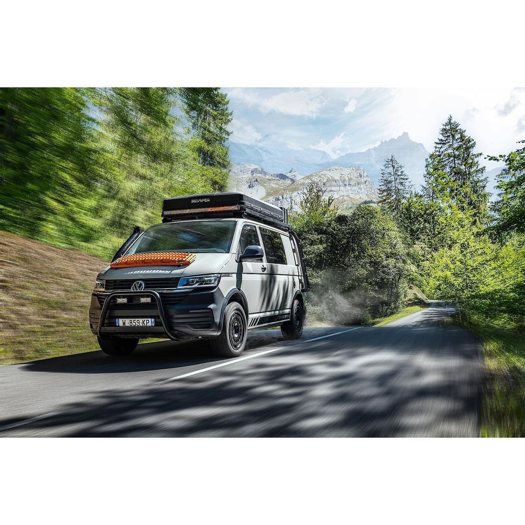 White and grey Volkswagen van with rooftop tent drives down a mountain road surrounded by green trees and mountains under a blue sky.