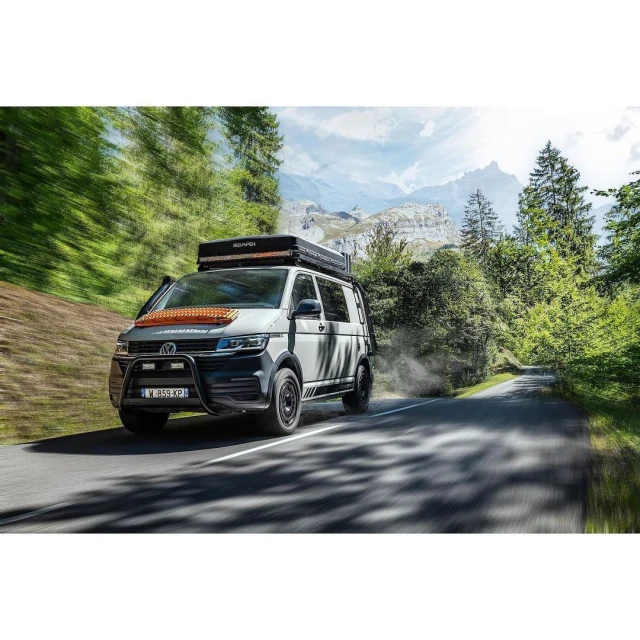 White and grey Volkswagen van with rooftop tent drives down a mountain road surrounded by green trees and mountains under a blue sky.