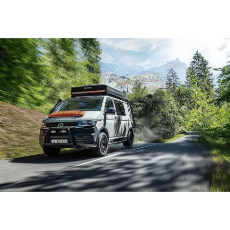 White and grey Volkswagen van with rooftop tent drives down a mountain road surrounded by green trees and mountains under a blue sky.