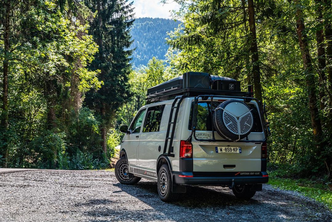 Off-white camper van with roof rack and spare tire parked on a gravel road in a lush forest with mountains in the distance.