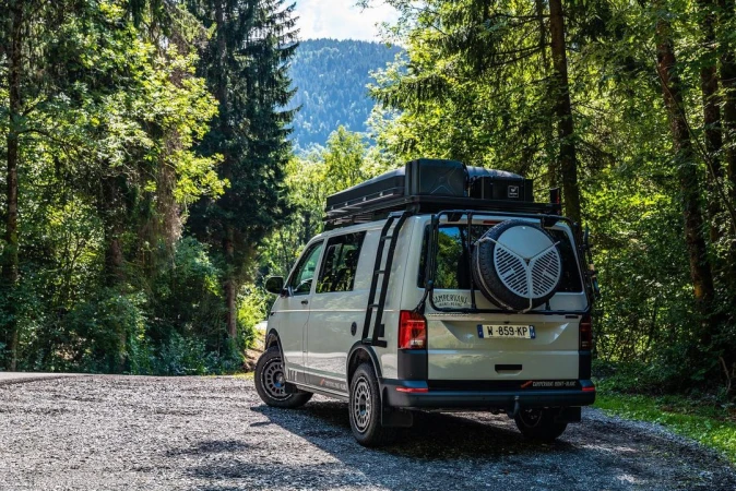 Off-white camper van with roof rack and spare tire parked on a gravel road in a lush forest with mountains in the distance.