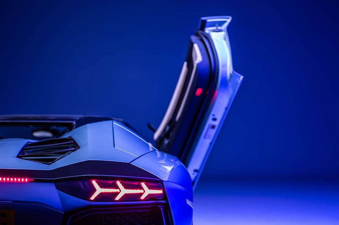Rear view of a blue Lamborghini with an open door against a gradient blue background, showcasing automotive design and luxury.