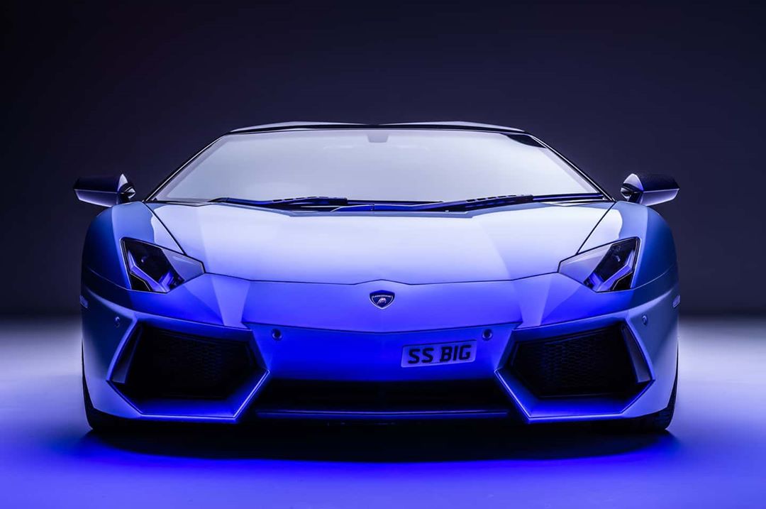 Front view of a blue Lamborghini Aventador with a license plate visible in a studio setting with dark background and blue lighting.