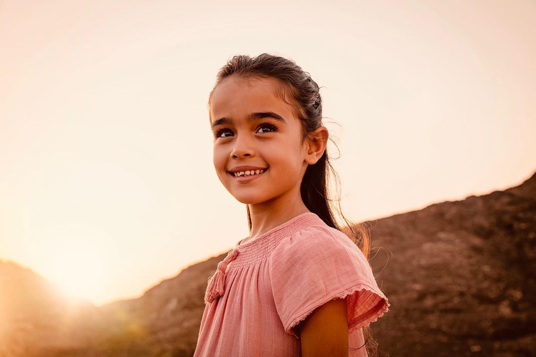 Young smiling child wearing a pink textured top outdoors against a hazy mountain backdrop during sunset.
