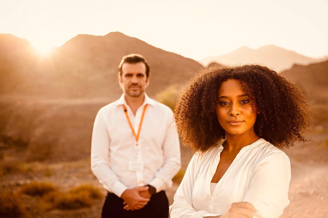 Portrait of a stylish woman with afro hairstyle, posing with confidence outdoors, accompanied by a male figure in the background.