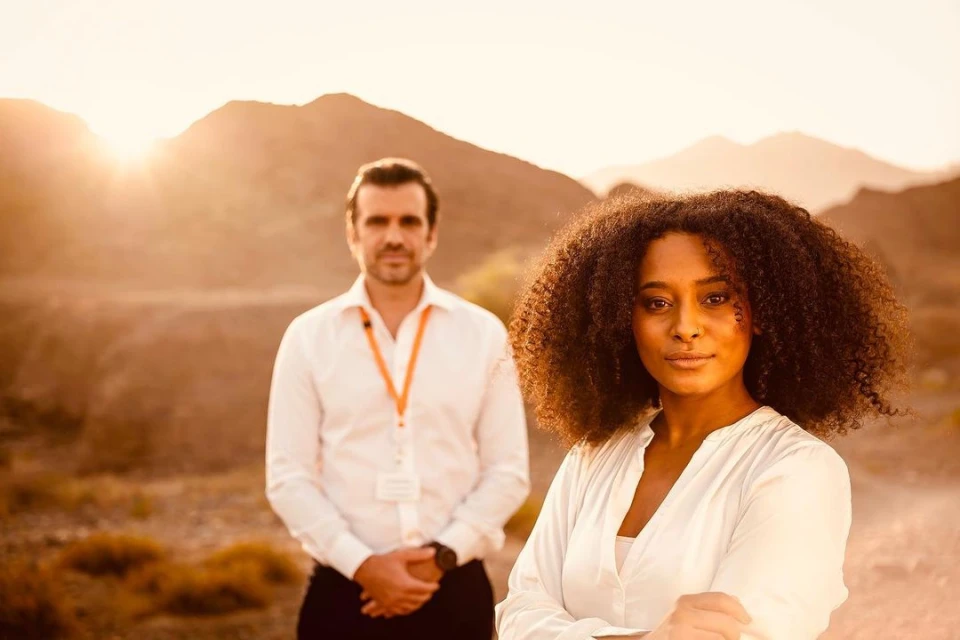 Portrait of a stylish woman with afro hairstyle, posing with confidence outdoors, accompanied by a male figure in the background.