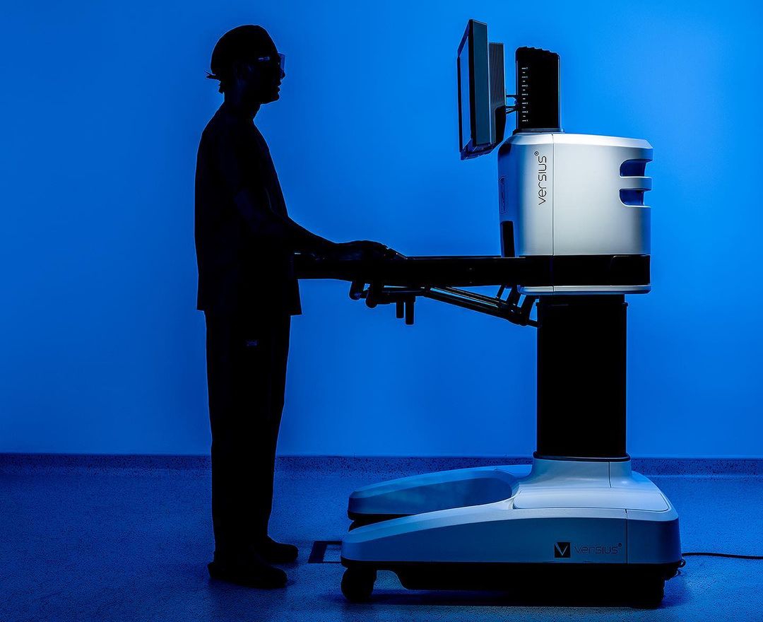 A surgical robot is used by a doctor silhouette, standing on a tiled floor in front of a gradient blue background in an indoor environment.