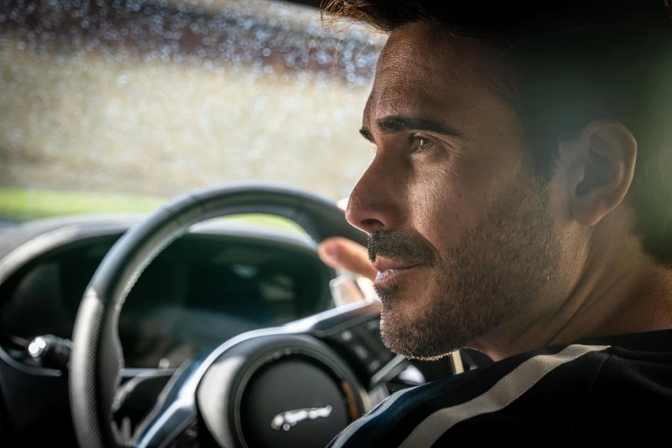 Adult male driving a black Jaguar car with a black steering wheel, captured in a close-up shot showcasing the interior of the vehicle.