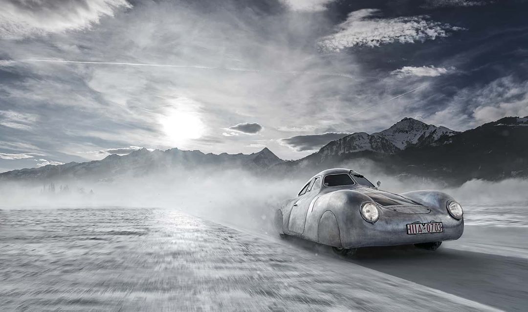 Vintage silver race car speeding across a snow-covered surface with mountains and a cloudy sky in the background, motion blur visible.