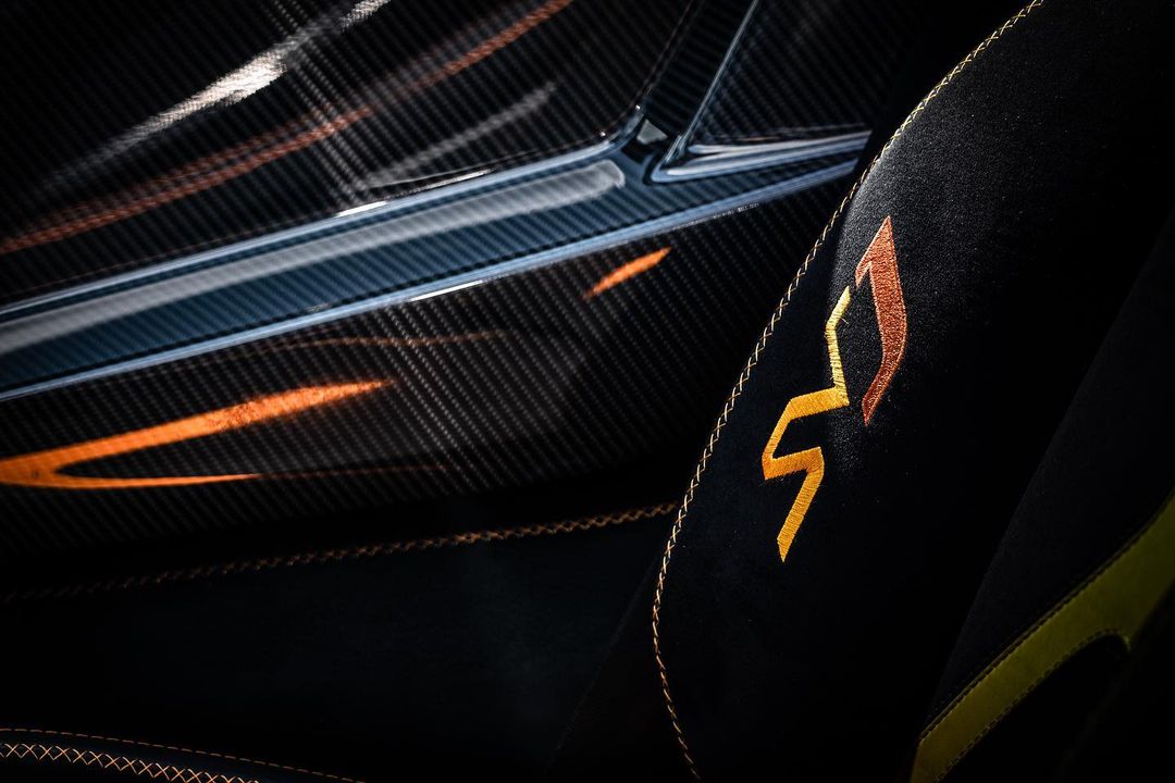 Close-up of a car interior showing a carbon fiber panel and a black suede seat with golden stitching and a golden emblem.