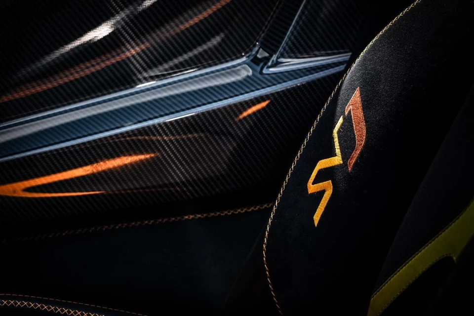 Close-up of a car interior showing a carbon fiber panel and a black suede seat with golden stitching and a golden emblem.