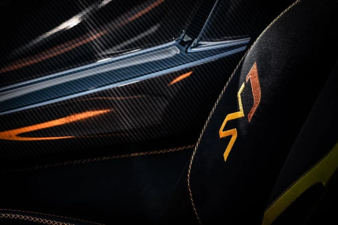 Close-up of a car interior showing a carbon fiber panel and a black suede seat with golden stitching and a golden emblem.