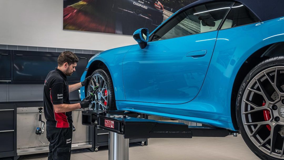 A mechanic working on the tire of a blue Porsche 911 on a vehicle lift inside a car service shop, with modern interiors and equipment.