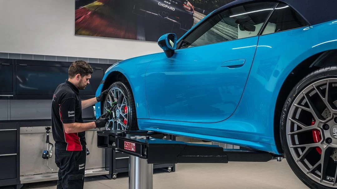 A mechanic working on the tire of a blue Porsche 911 on a vehicle lift inside a car service shop, with modern interiors and equipment.