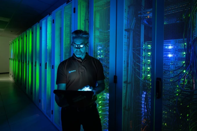 Technician wearing glasses and polo shirt using tablet inside server room with glowing blue and green server racks