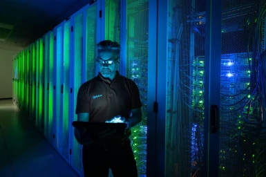 Technician wearing glasses and polo shirt using tablet inside server room with glowing blue and green server racks