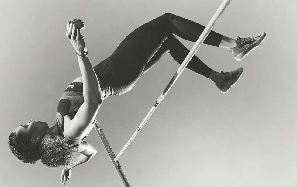 Monochromatic shot of an adult female high jumper in mid-air, arched over the bar, against a bright sky. Athletic apparel visible.
