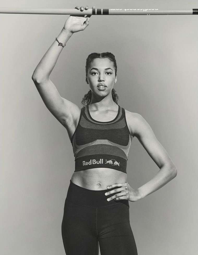 Female athlete stands confidently holding a pole vault bar in a studio, wearing a Red Bull crop top and athletic leggings against a grey backdrop.