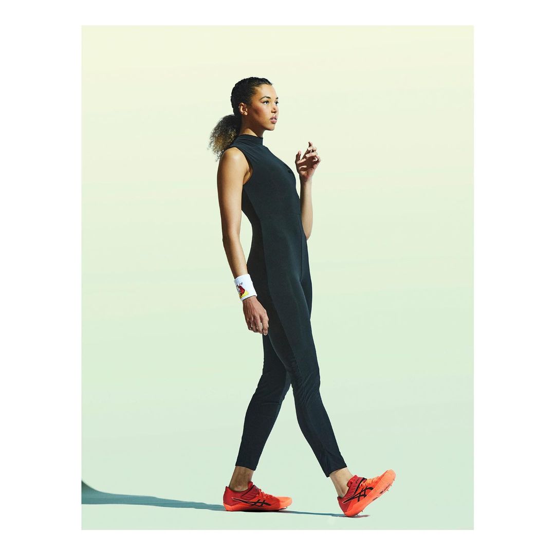 Athletic woman in black jumpsuit walking in a studio, wearing orange sneakers and a wristband, against a gradient background.