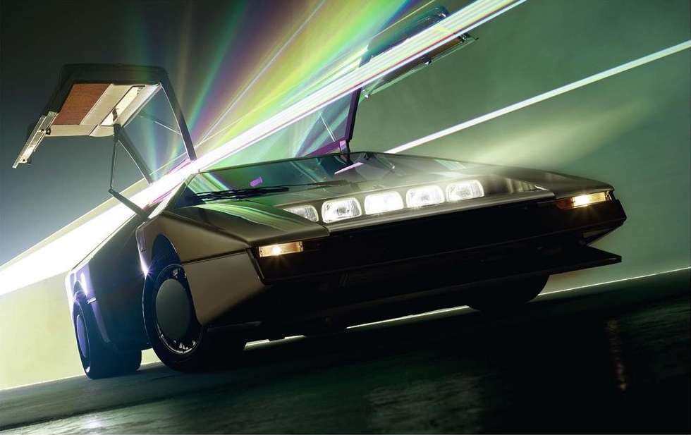 Sleek silver sports car with gull-wing doors open, illuminated by bright, multicolored lights in a dark, indoor setting.