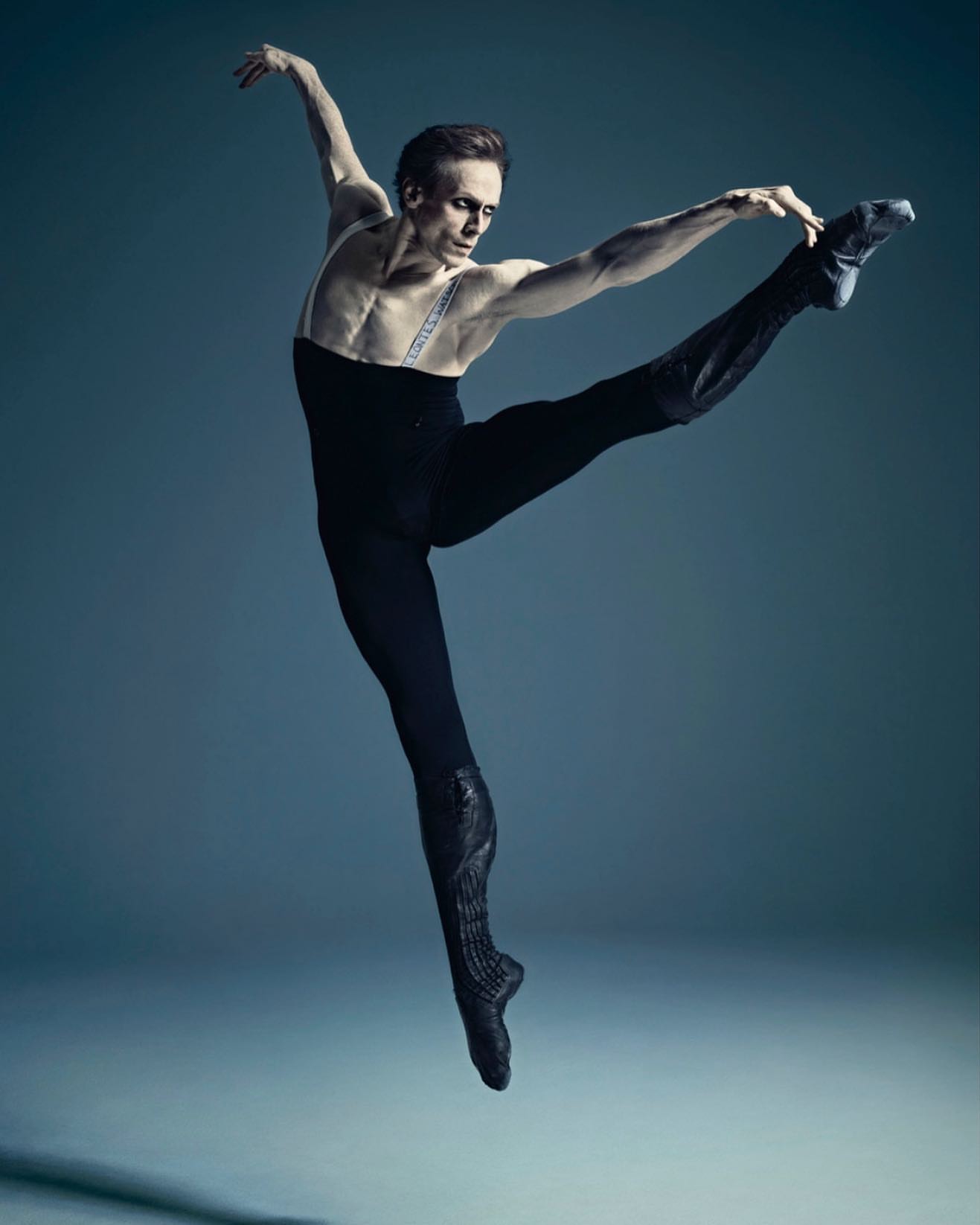 Male ballet dancer in mid-air, performing a leap in a black costume against a blue background, showcasing athleticism and artistry.