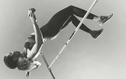 Black female athlete clearing the high jump bar mid-air, captured in a dynamic sports action shot by Rick Guest.