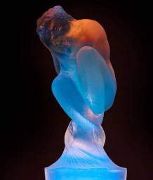 A translucent glass sculpture of a human figure curled tightly, illuminated by blue and warm orange light against a dark background.