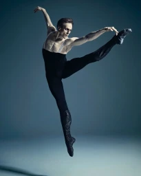 Male ballet dancer mid-air leaping in a high-extension pose wearing black tights and soft leather boots against a solid grey background.