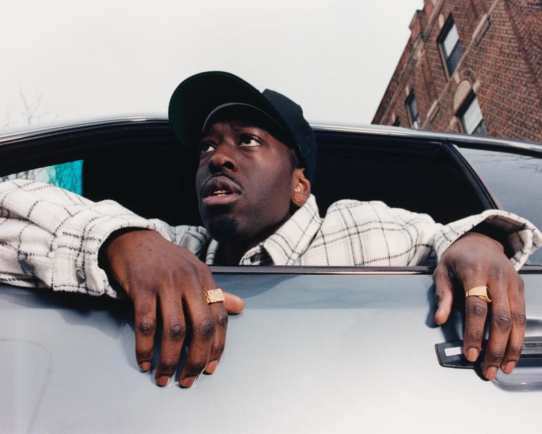 Portrait of a dark-skinned man in car, wearing baseball cap and gold rings, gazing upwards on an overcast day in an urban setting.